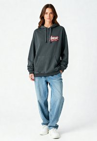 Donna in piedi che indossa un oversize grigio scuro con la scritta "Amour", jeans larghi blu chiaro e sneakers bianche su uno sfondo bianco.