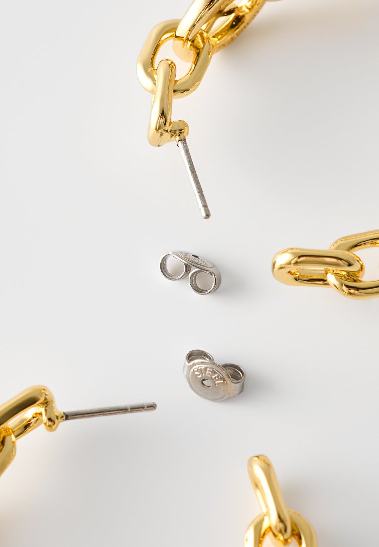 Gouden schakels met een glanzende afwerking, zilveren oorbel achterop en een stalen cirkelsteun op een witte ondergrond. Texturen en kleuren contrasteren duidelijk.