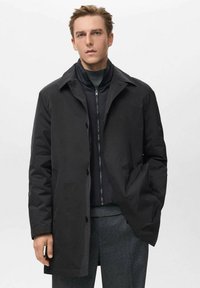 Abrigo negro largo con cuello tipo camisa, botones en el frente y bolsillos laterales. Llevado sobre una chaqueta negra con cremallera y pantalones grises. Textura suave.