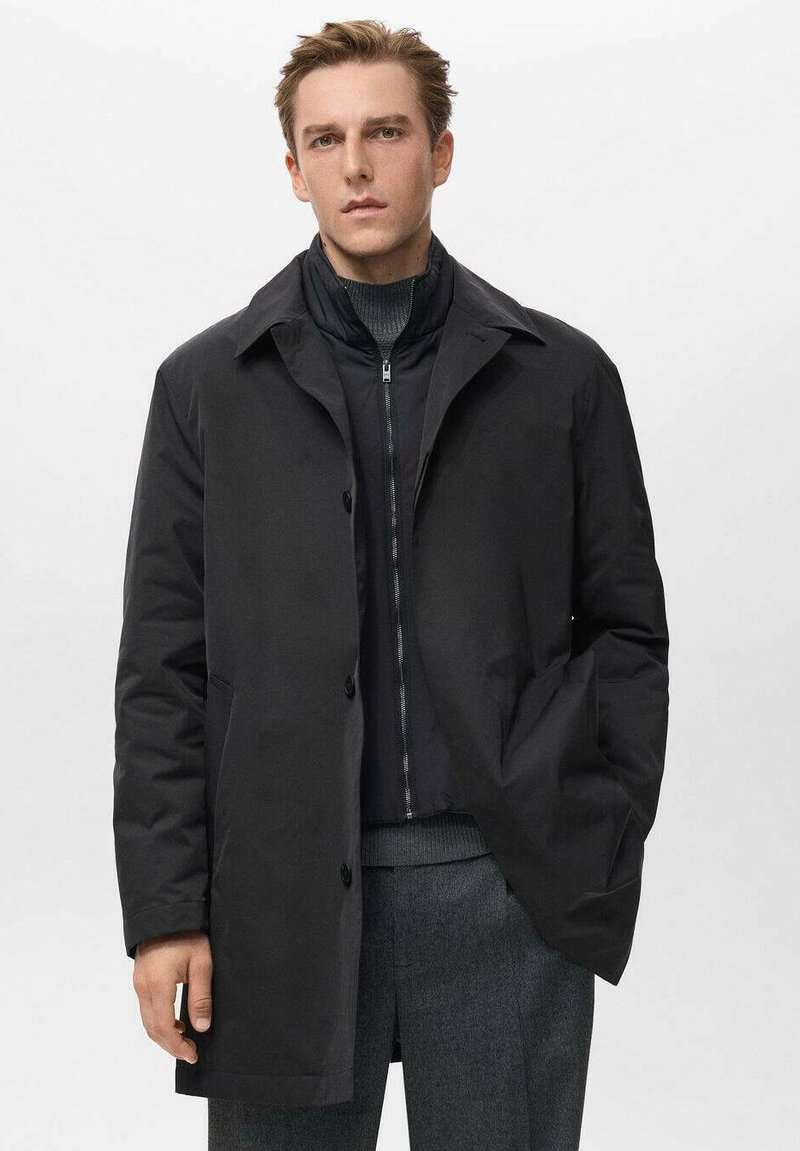 Abrigo negro largo con cuello tipo camisa, botones en el frente y bolsillos laterales. Llevado sobre una chaqueta negra con cremallera y pantalones grises. Textura suave.