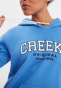 Sudadera azul con cordones blancos, con la frase "CREEK ORIGINAL Desde 1974" impresa en blanco y azul marino. Textura de tela suave, diseño casual.
