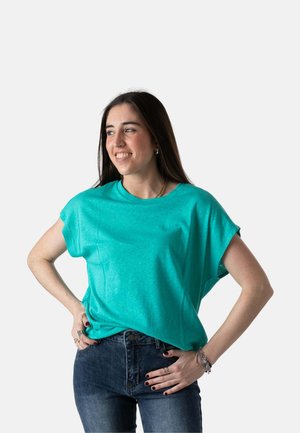 Joven mujer con camiseta de manga corta turquesa y vaqueros, sonriendo y mirando hacia su izquierda con las manos en las caderas contra un fondo liso.