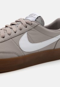 Pilki odiniai sportbačiai su balta Nike swoosh, apvaliu pirštu ir ruda guma. Šnerves yra šviesiai pilkos spalvos, su lygiu paviršiumi.