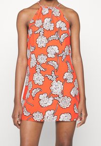 Femme portant une robe orange sans manches à col haut avec un grand motif floral blanc, debout les bras détendus le long du corps.