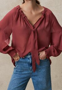 Blusa trasparente mogano con chiusura a nastro e polsini arricciati, abbinata a jeans in denim blu a vita alta. Tessuto leggero e fluido.