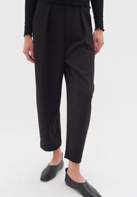 Pantaloni tailored neri con pieghe, texture liscia, lunghezza cropped e dettagli con spacco laterale. Abbinati a scarpe nere slip-on.