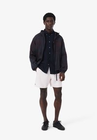 Giacca leggera da uomo di un blu scuro con cappuccio, abbinata a una camicia button-up blu scuro, pantaloni corti di un rosa pallido e sneakers nere. Texture lisce visibili.