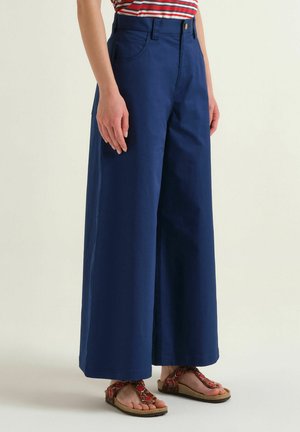 Person trägt hoch taillierte, weit geschnittene marineblaue Hose, rot gestreiftes Hemd und rote gemusterte Sandalen, steht vor einem einfarbigen hellen Hintergrund.