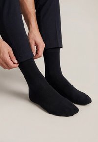 Muškarac obučen u crne čarape i tamne hlače prilagođava rub hlača na jednobojnoj svijetloj pozadini.