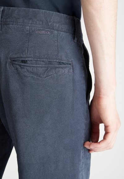 Ciemnoniebieskie spodnie wykonane z teksturowanej tkaniny, z tylną kieszenią z delikatnym przeszyciem i metką z logo przy pasku. Minimalistyczny design.