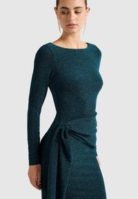Šaty s dlhým rukávom v tyrkysovej farbe, vyrobené z textúrovaného, trblietavého materiálu. Majú priliehavý strih s uzlovým detailom v páse.