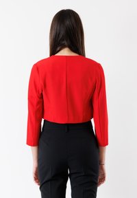 Giacca rossa corta con maniche a tre quarti, tessuto liscio, collo alto e dettaglio della cucitura posteriore, abbinata a pantaloni neri.