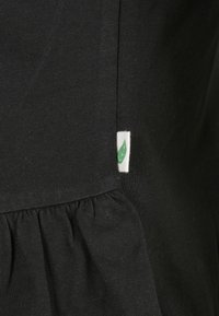 Svart bomullstop med samlad nederkant. Har en liten vit etikett med en grön löv-grafik. Mjuk textur, enkel design och skräddarsydd passform.