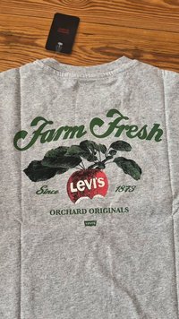 Grå skjorte med grønn tekst "Farm Fresh" og "Orchard Originals", et rødt eple med "Levi's" logo, og et svart Levi's merke på en treoverflate.
