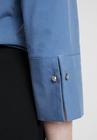 Lichtblauwe shirtmouw met een gespleten manchet, voorzien van decoratieve hardware in zilverkleur bij de opening, gemaakt van gladde stof.