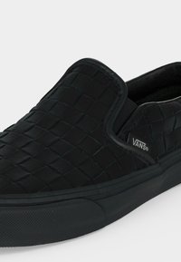 Μαύρο παπούτσι Vans τύπου slip-on με υφαντό υφή στην εξωτερική επιφάνεια και λαστιχένια σόλα, με ελαστικές πλευρικές λεπτομέρειες και μικρό λογότυπο Vans στη πλευρά.