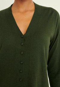 Cardigan verde con scollo a V, caratterizzato da sette bottoni neri sul davanti. Tessuto a maglia morbido e design aderente.
