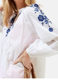 Blusa bianca con ricami floreali blu sulle maniche, polsini elastici e vestibilità comoda. Il materiale sembra leggero e testurizzato.