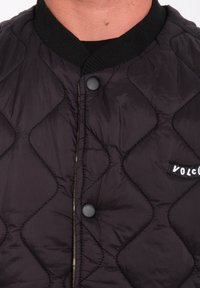 Chaqueta acolchada negra con cuello de canalé, botones negros y un pequeño logo "VOLCOM" en el frente. Tela lisa y ligera.