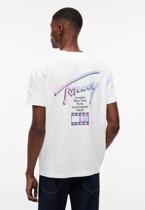 Fehér T-shirtöt viselő férfi, rajta pasztell színű "Tommy" felirat, városnevek és a Tommy Jeans logó a hátoldalon, sötétkék farmerral párosítva.