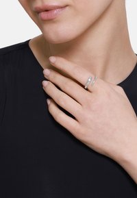 Anello in argento con una finitura liscia e lucida. Presenta due bande curvate, una con texture e l'altra liscia, appoggiate su una mano su uno sfondo nero.