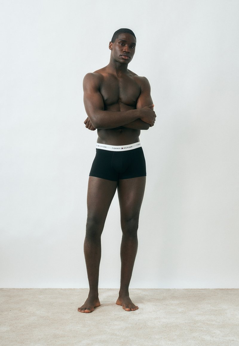 Μυώδης άνδρας όρθιος ξυπόλητος πάνω σε χαλί, φοράει μαύρα μποξεράκια Tommy Hilfiger, με σταυρωμένα τα χέρια, μπροστά σε απλό ανοιχτό γκρι φόντο.