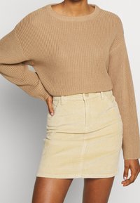 Pull beige en maille avec un col rond et des manches longues, associé à une mini-jupe en velours côtelé jaune clair avec des poches avant et une taille ajustée.