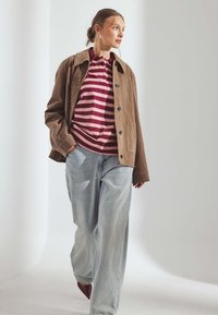 Donna che cammina con jeans larghi color azzurro chiaro, giacca marrone con bottoni e camicia a righe rosse su sfondo chiaro uniforme.