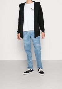 Svart stickad huva cardigan över en vit t-shirt, tillsammans med ljusblå denimjeans och svarta sneakers med vita sulor.