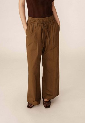 Pantalons larges marron avec une taille élastique et un cordon de serrage. Le tissu semble lisse et légèrement brillant. Associés avec des talons tissés sombres.