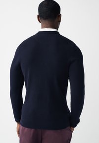 Navnøje strikket sweater med et tekstureret mønster, rund hals og tætsiddende ærmeafslutninger. Båret med mørke bukser, der har et glat stof og skræddersyet pasform.