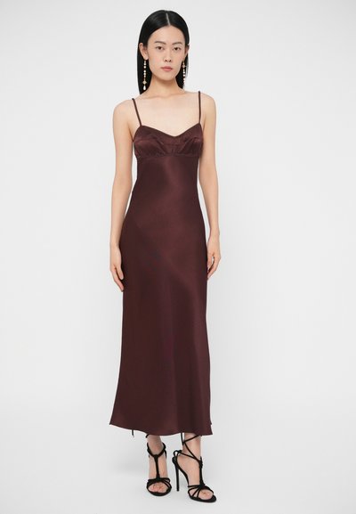 Žena v dlhých tmavo bordových saténových šatách bez rukávov s tenkými ramienkami a čiernymi sandálmi na vysokom opätku, stojaca pred jednoliatym pozadím.