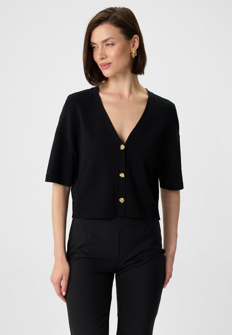 Schwarze Strickjacke mit V-Ausschnitt, kurzen Ärmeln und cropped Saum. Mit drei goldenen Knöpfen vorne und einer strukturierten Oberfläche.