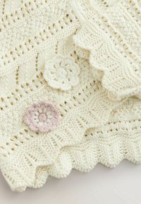 Tissu tricoté couleur crème avec des bords festonnés, présentant des motifs de points détaillés et deux petites applications de fleurs au crochet, une blanche et une rose.