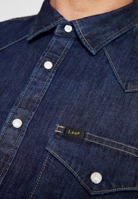 Granatowa koszula dżinsowa z kołnierzykiem w kształcie szpicu, białymi guzikami, kontrastującym żółtym przeszyciem i małą czarną metką ze złotym tekstem.