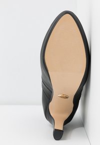 Chaussure à talon en cuir noir à bout pointu avec semelle lisse couleur tan et embout de talon noir texturé, posée verticalement contre des murs blancs.