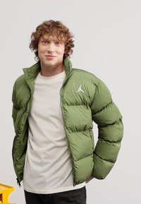 Giacca verde gonfiabile con zip frontale, caratterizzata da sezioni trapuntate orizzontali e un logo bianco sul lato sinistro del petto. Indossata sopra una camicia color crema.