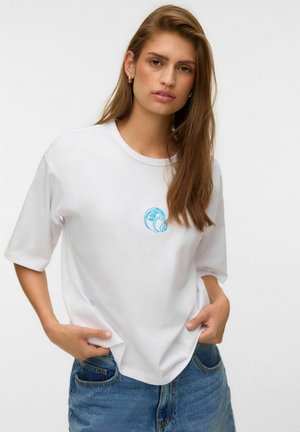 Ung kvinde med langt brunt hår iført en hvid t-shirt med et lille blåt muslingemotiv og blå jeans, hænderne i lommerne.
