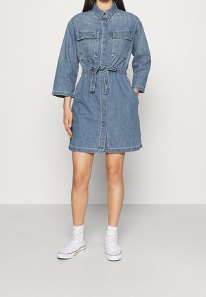 Femme portant une robe-chemise en jean bleu avec manches longues, ceinture à la taille, deux poches poitrine, chaussettes blanches et baskets blanches montantes, debout sur un sol gris.
