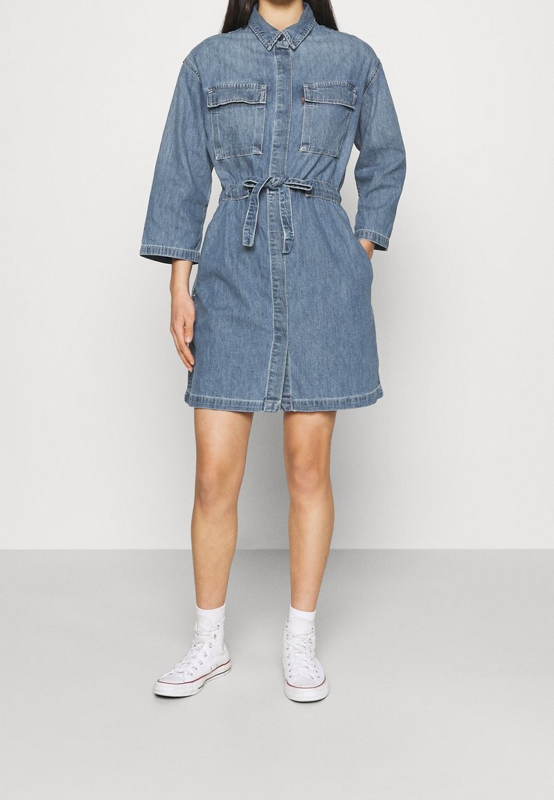 Femme portant une robe-chemise en jean bleu avec manches longues, ceinture à la taille, deux poches poitrine, chaussettes blanches et baskets blanches montantes, debout sur un sol gris.