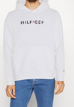 Felpa con cappuccio bianca con la scritta "HILFIGER" in colori misti sul petto, tasca anteriore, indossata con jeans blu.