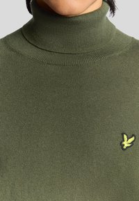 Olivgrön polotröja med ribbad textur och figursydd design, med en liten gul broderad logotyp av en fågel på bröstet.