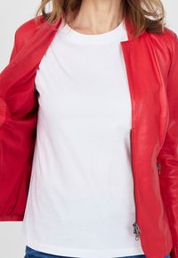 Červená kožená bunda rozepnutá přes bílý tričko, která zdůrazňuje hladkou texturu a elegantní design bundy. Výstřih je kulatý a ležérní.