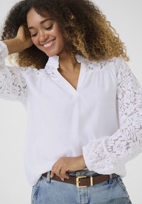 White blouse with lace detail on sleeves, v-neckline, and gathered accents. Paired with high-waisted blue jeans and a brown belt.