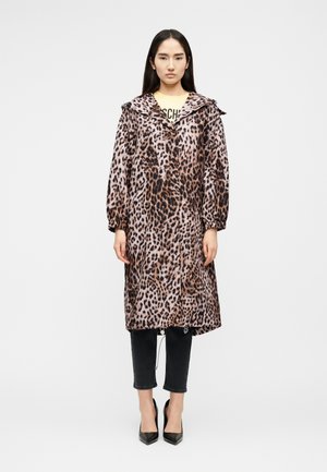 Γυναίκα όρθια, φοράει ένα μακρύ παλτό με λεοπάρ τύπωμα, μαύρο παντελόνι μέχρι τον αστράγαλο και μαύρες μυτερές ψηλοτάκουνες γόβες σε απλό φόντο.