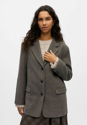 Donkergrijze wollen blazer met een normale kraag, twee voorkruiszakken en drie knopen. Gelaagd over een lichtbeige sweater.