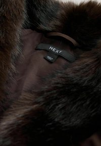Pruun faux karusnaha krae, mis on kinnitatud siledale, tumedale kangale. Omab sildiga "NEXT" ja "MADE IN CHINA".