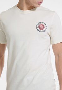 Krēmkrāsas kokvilnas T-krekls ar apaļu kakla izgriezumu un īsām piedurknēm; kreisajā krūtīs ir apļa sarkans un melns logo.