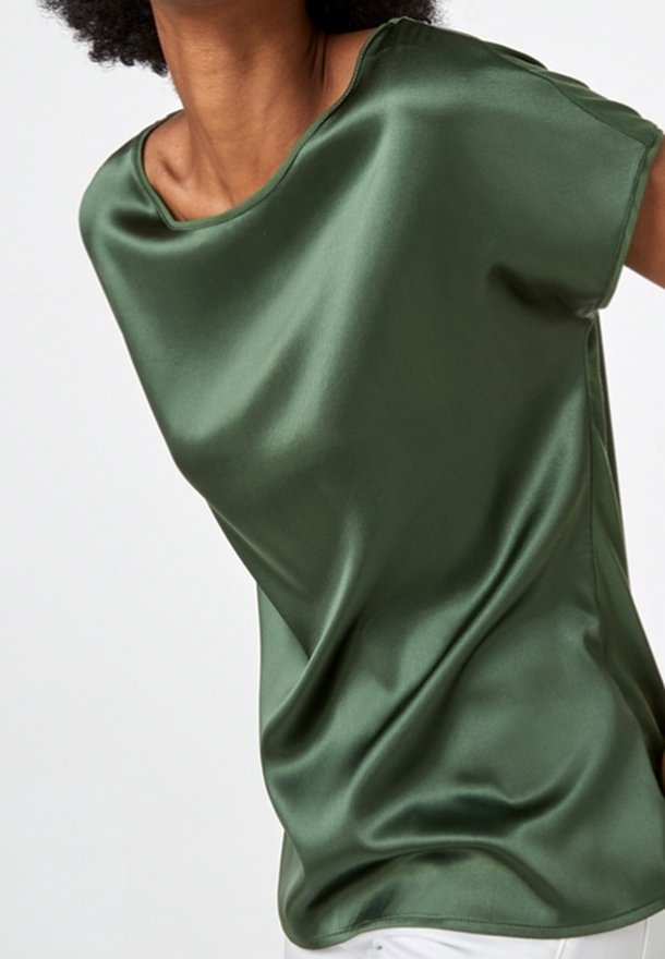 Femme portant un chemisier en satin olive brillant, sans manches, à coupe ample, sur un fond blanc uni.