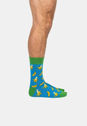 Gambe con calzini di un blu brillante con risvolti e punte verdi, decorati con illustrazioni di banane gialle e banane sbucciate.